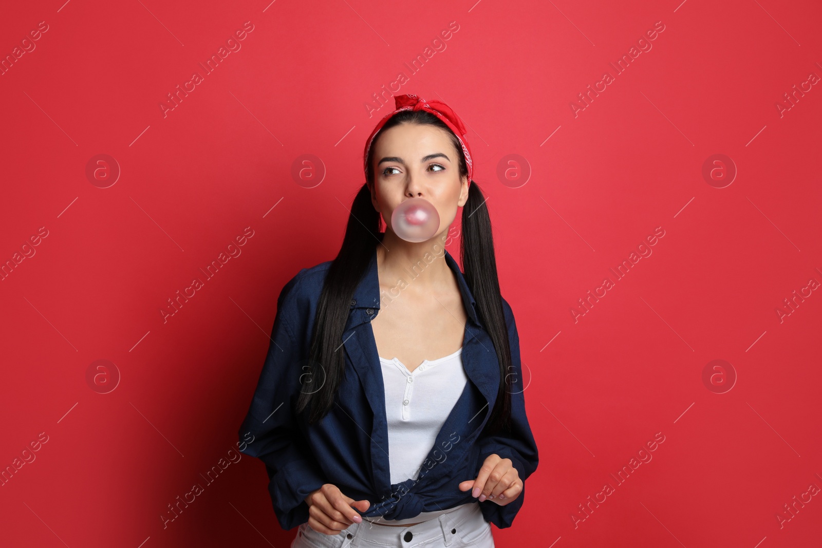 Photo of Fashionable young woman in pin up outfit blowing bubblegum on red background