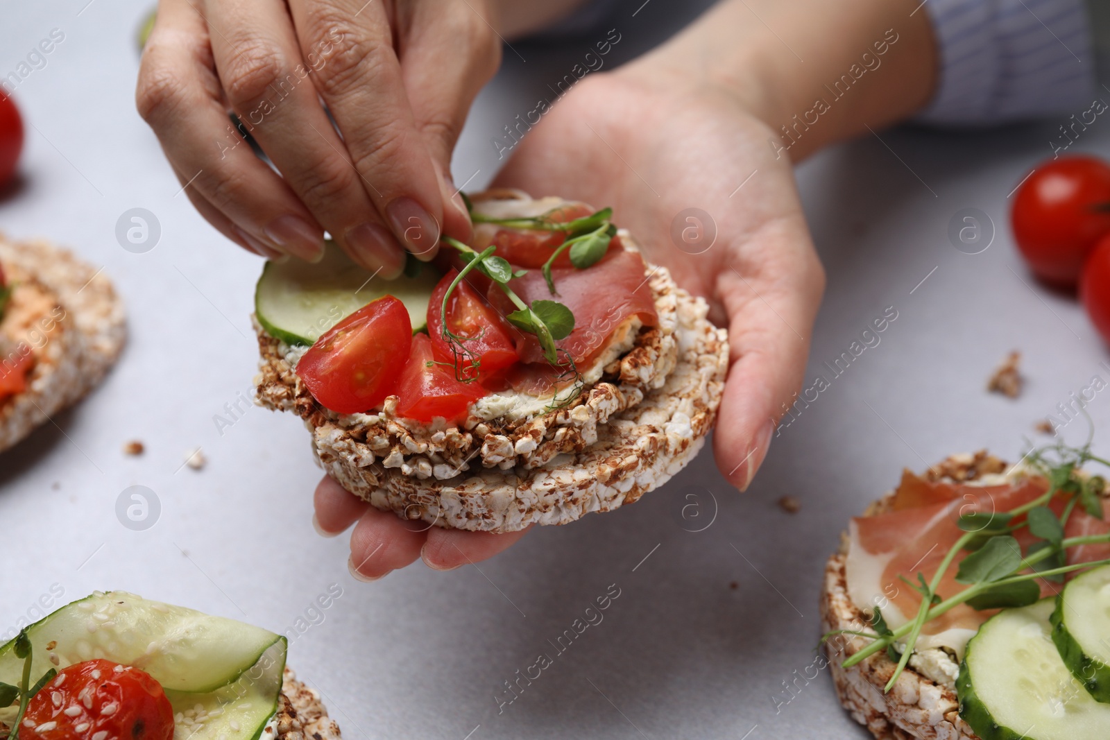 Photo of Woman adding microgreens onto crunchy buckwheat cakes with prosciutto, pieces of tomato and cucumber slice at white table, closeup