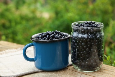 Photo of Jar and cup of delicious bilberries on wooden table outdoors
