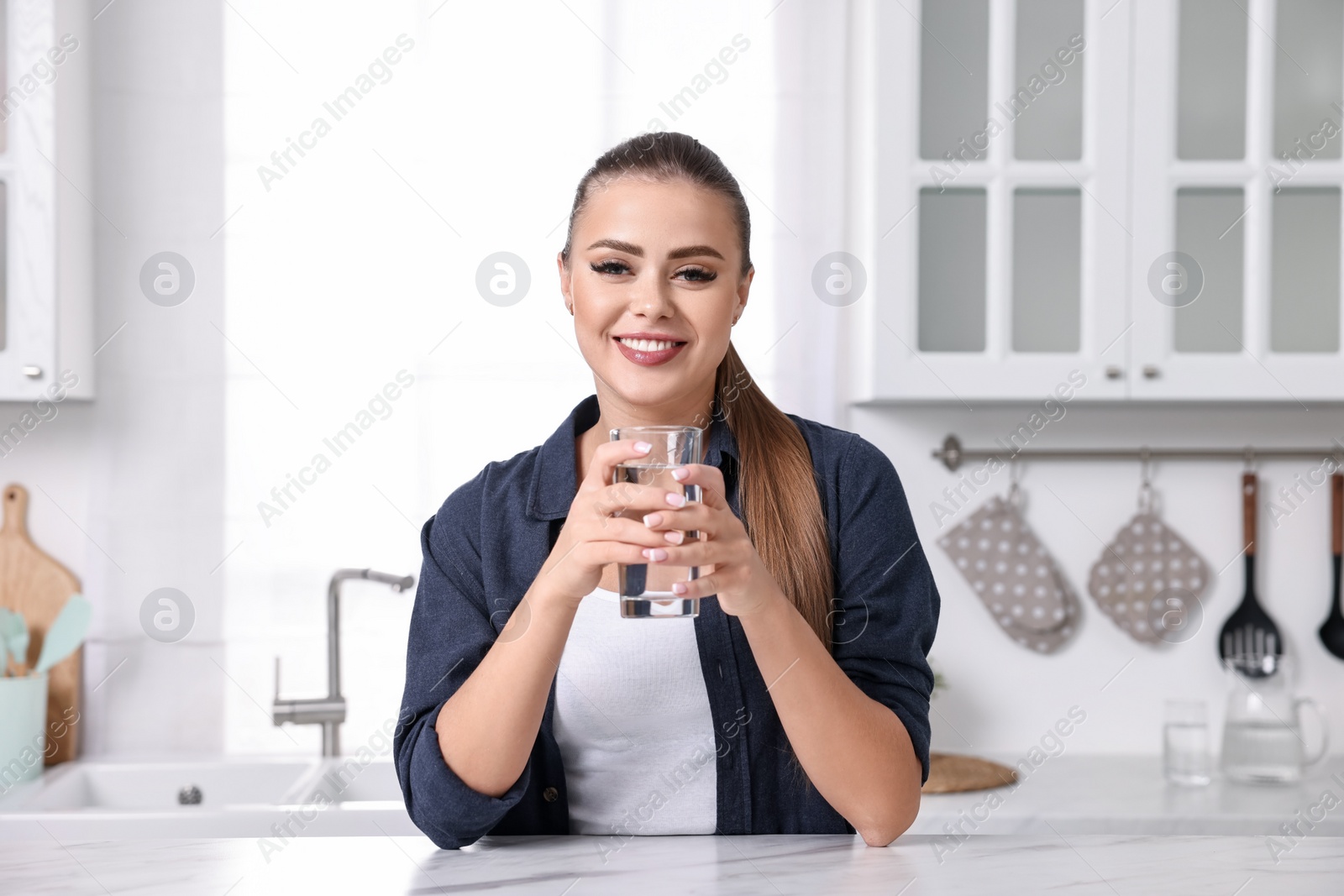 Photo of Happy woman with glass of fresh water at white marble table in kitchen