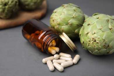 Photo of Bottle with pills and fresh artichokes on grey wooden table, closeup