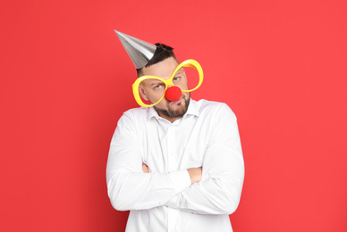 Emotional man with large glasses, party hat and clown nose on red background. April fool's day