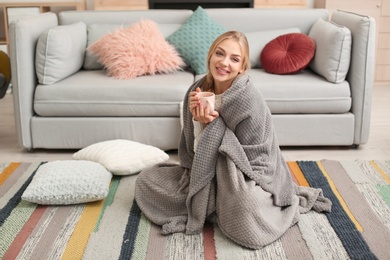 Photo of Beautiful young woman wrapped in plaid sitting with cup of coffee on floor at home. Winter atmosphere