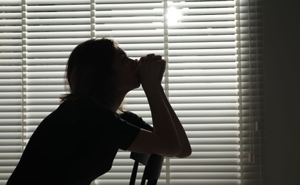 Photo of Silhouette of sad young woman near closed blinds indoors, space for text