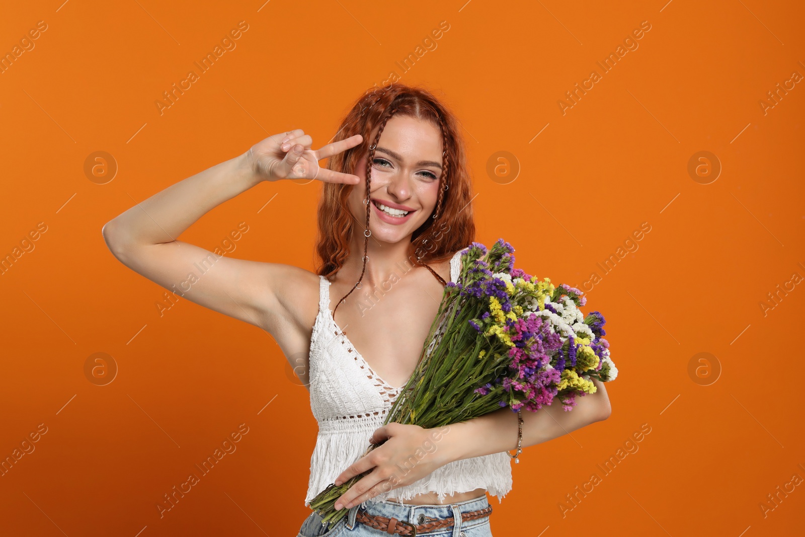 Photo of Beautiful young hippie woman with bouquet of colorful flowers showing V-sign on orange background