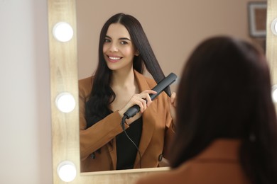 Photo of Beautiful happy woman using hair iron near mirror indoors
