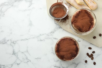 Photo of Delicious tiramisu in glasses, cookies, coffee beans and sieve with cocoa powder on white marble table, top view. Space for text