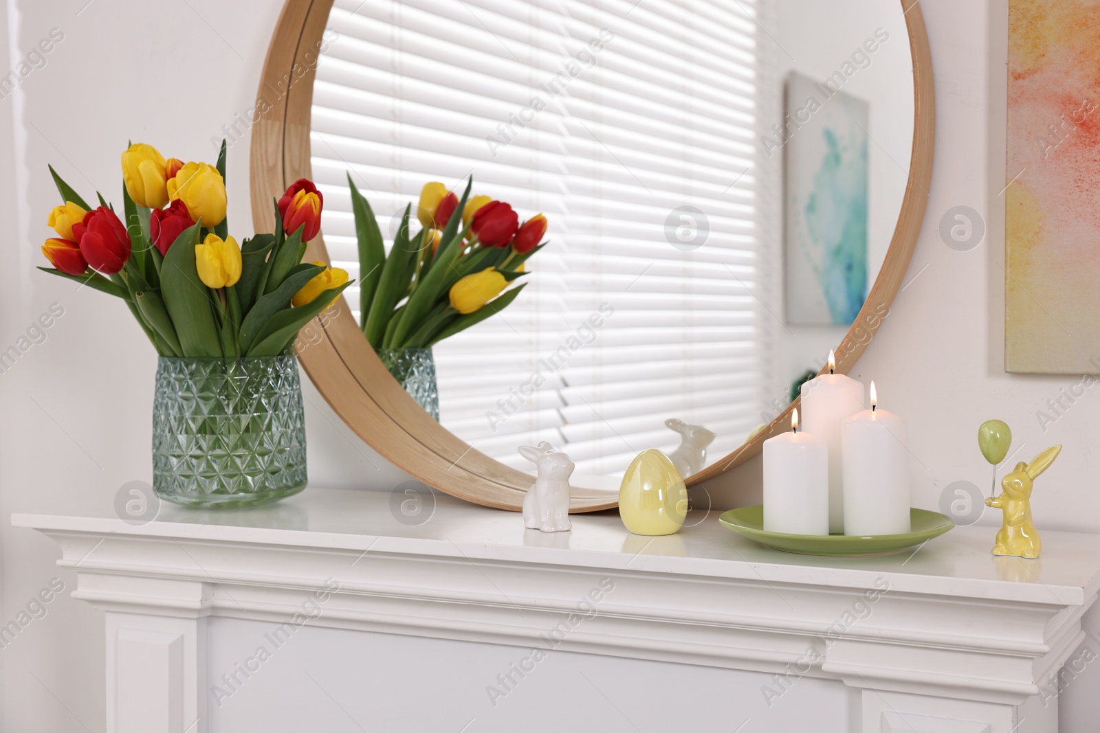 Photo of Easter decorations. Bouquet of tulips in vase, burning candles and bunny figures on fireplace at home