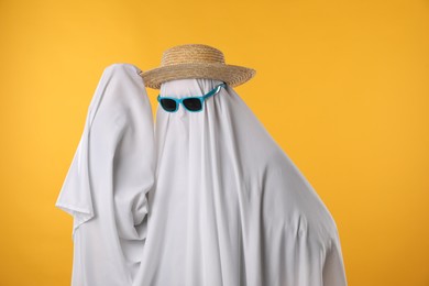 Person in ghost costume, sunglasses and straw hat on yellow background, space for text