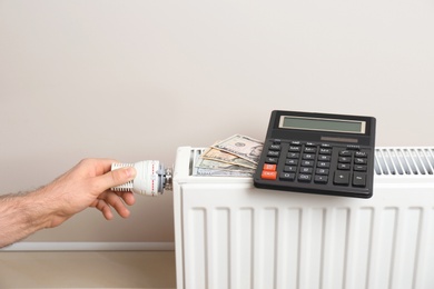 Man adjusting temperature of heating radiator with calculator and money on it indoors