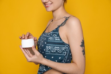 Woman with tattoo holding jar of cream against yellow background, closeup