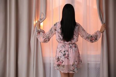 Young woman wearing floral print dress near window at home, view from back