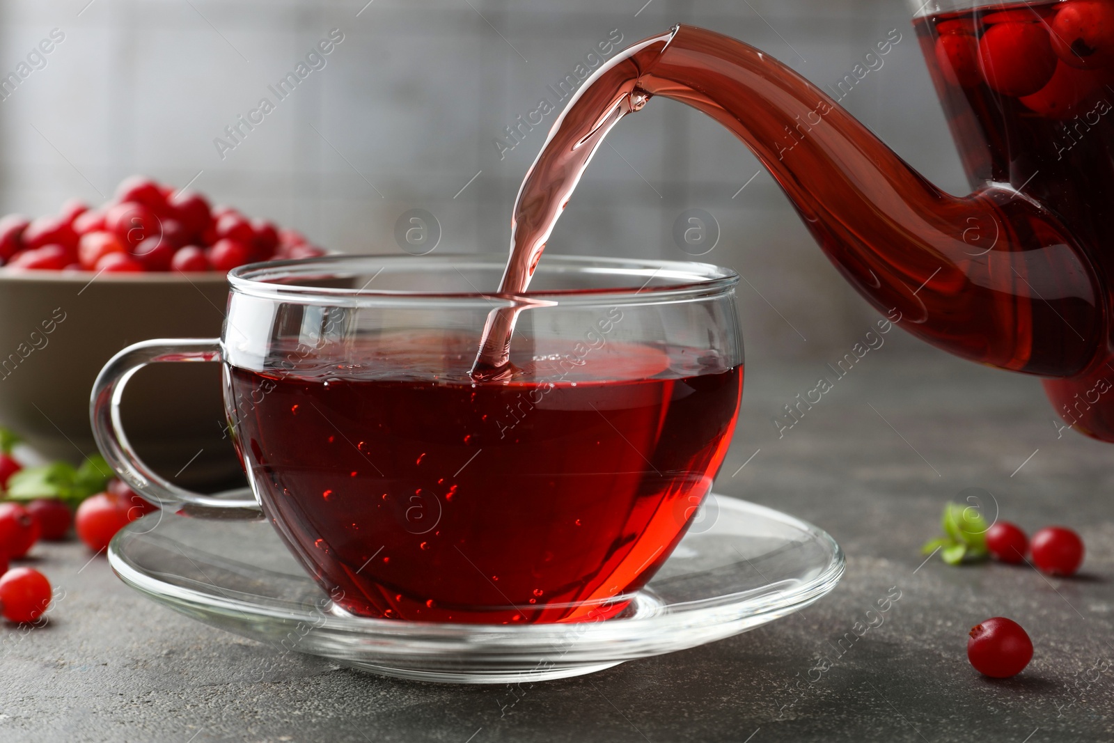 Photo of Pouring hot cranberry tea into glass cup and fresh berries on light grey textured table, closeup