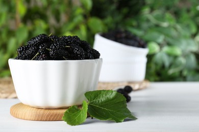 Fresh ripe black mulberries in bowl on white wooden table against blurred background. Space for text