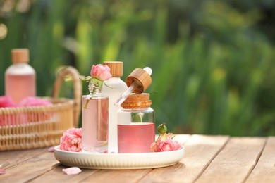 Photo of Bottles of rose essential oil and flowers on wooden table outdoors, space for text