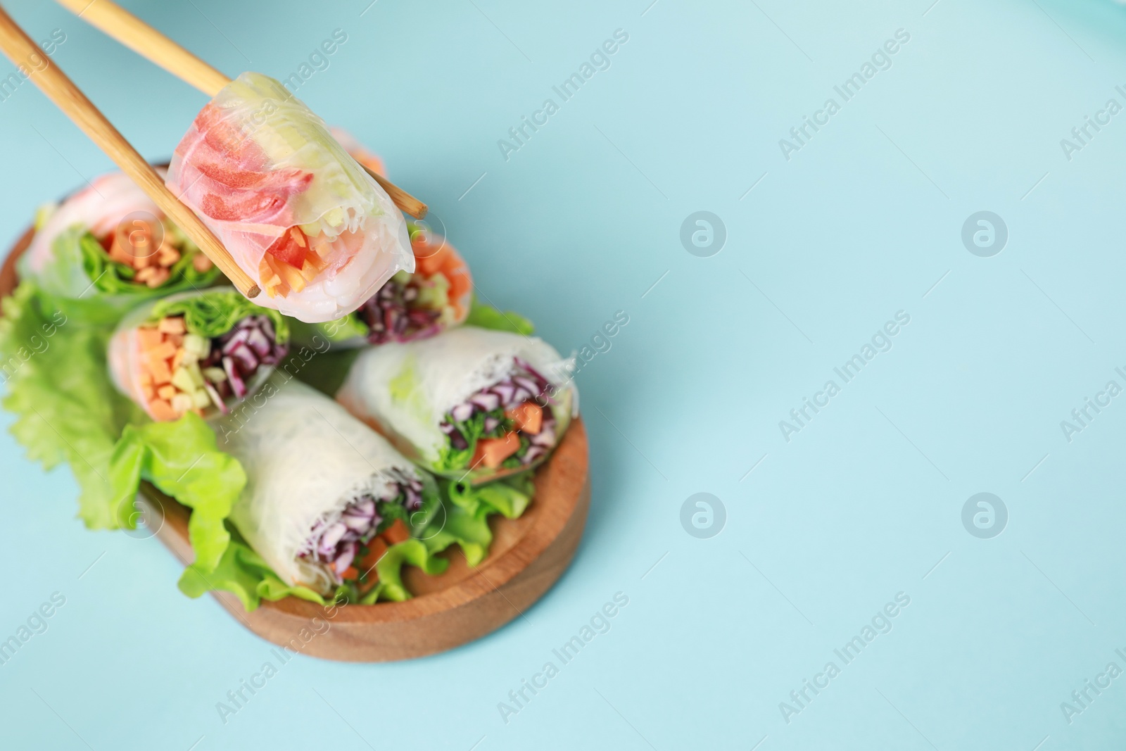 Photo of Holding delicious spring roll with chopsticks on light blue background, closeup. Space for text