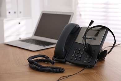 Photo of Stationary phone and headset on wooden table indoors. Hotline service