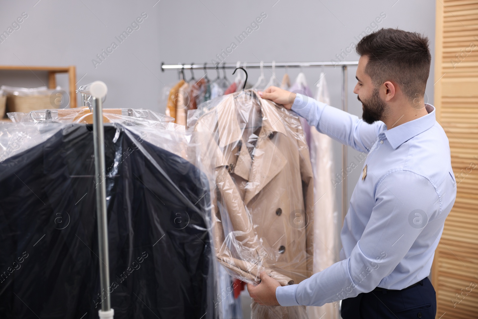 Photo of Dry-cleaning service. Worker holding hanger with coat in plastic bag near rack with clothes indoors