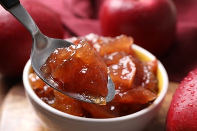 Photo of Spoon with delicious apple jam above bowl on table, closeup