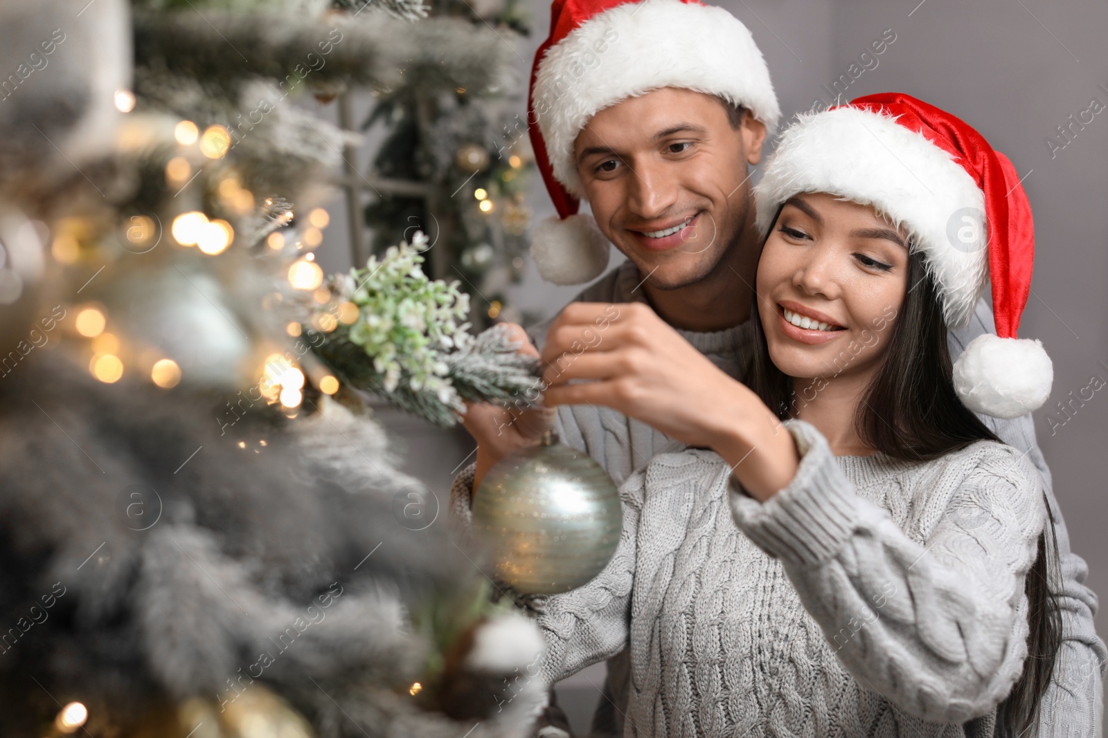 Photo of Happy young couple in Santa hats decorating Christmas tree together at home