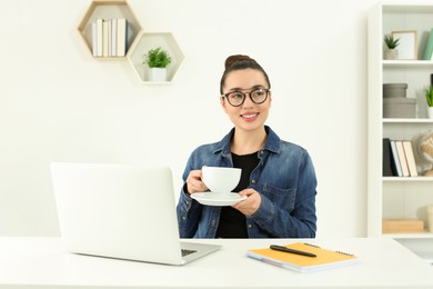 Photo of Home workplace. Happy woman with cup of hot drink near laptop at white desk in room