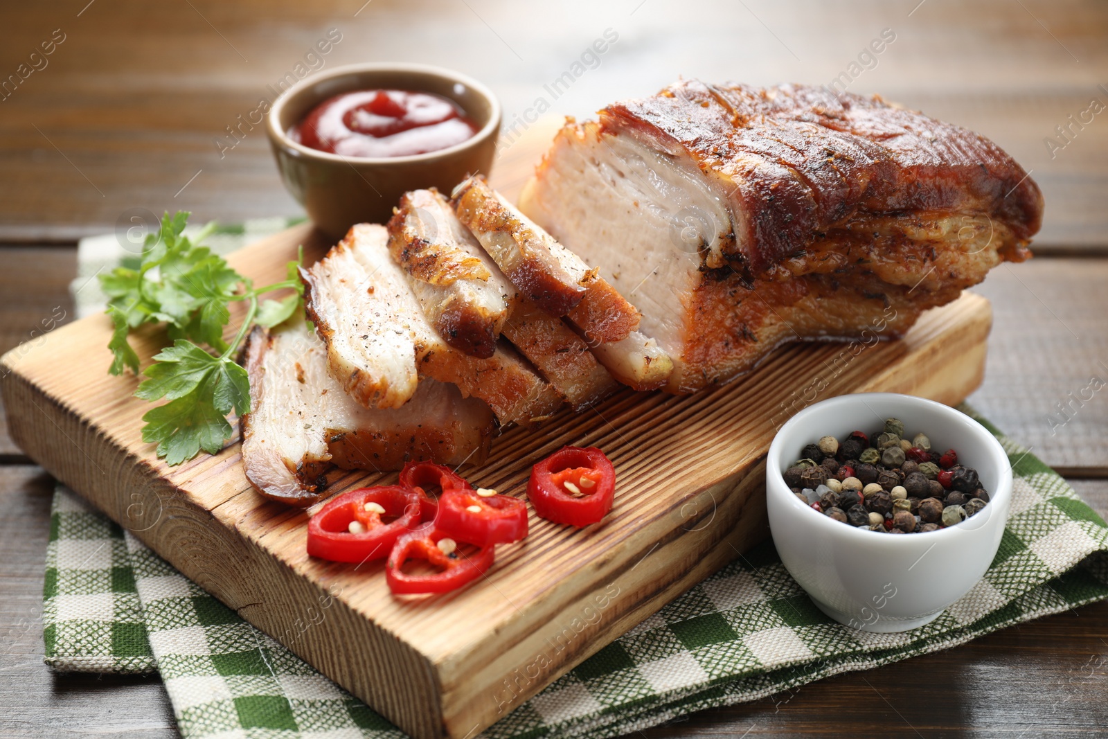 Photo of Pieces of baked pork belly served with sauce, chili pepper and parsley on wooden table, closeup