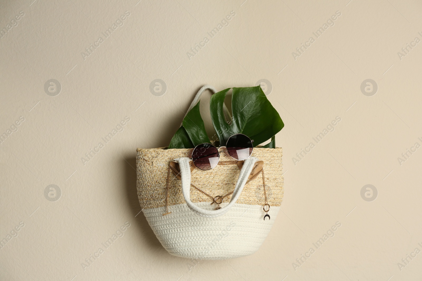 Photo of Elegant woman's straw bag with tropical leaf and accessories hanging on beige background