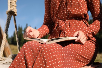 Photo of Young woman reading book on wooden swing outdoors, closeup