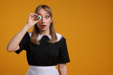 Photo of Emotional woman in scary maid costume with decorative eyeball on orange background, space for text. Halloween celebration