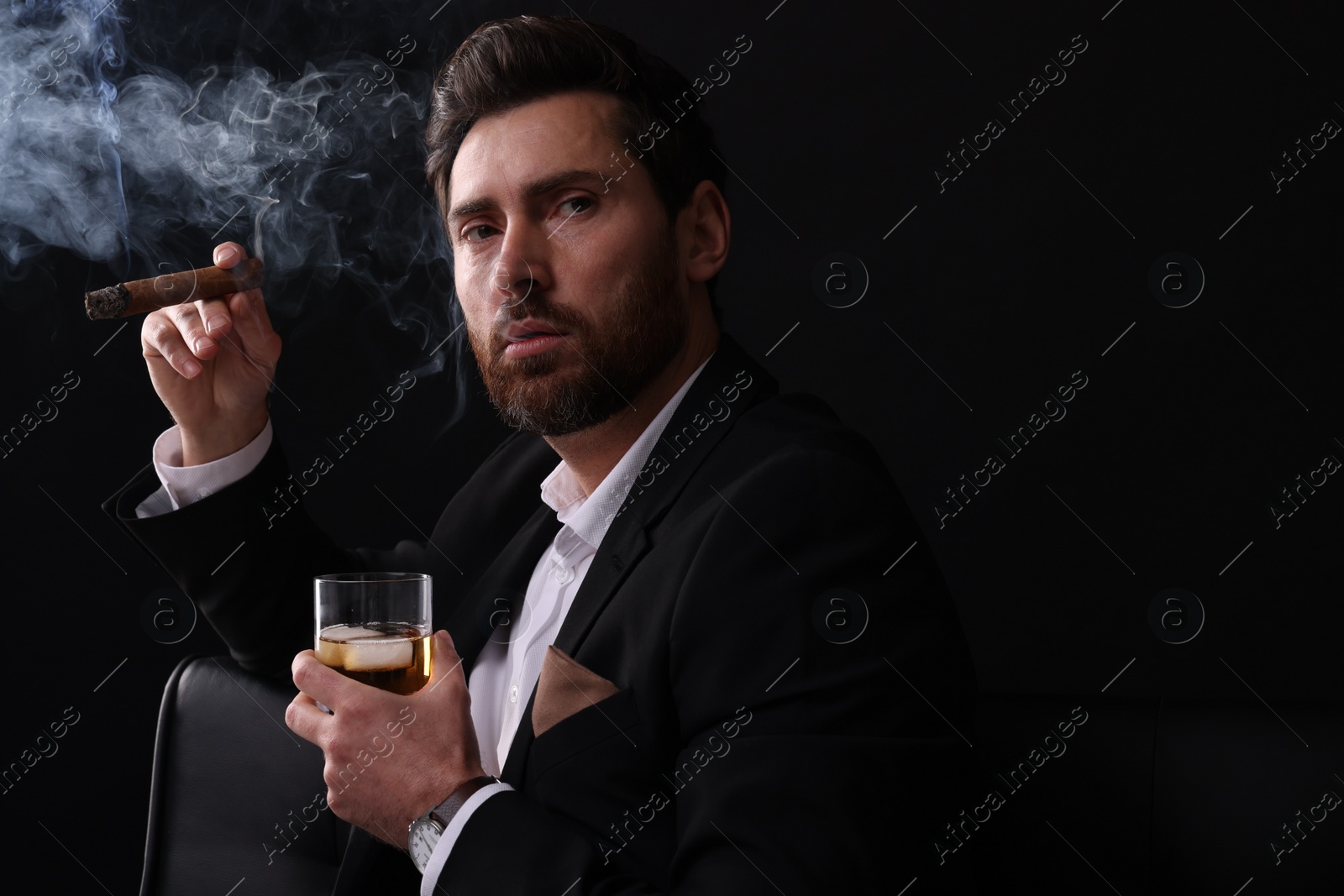 Photo of Handsome man in elegant suit with glass of whiskey smoking cigar on black background. Space for text