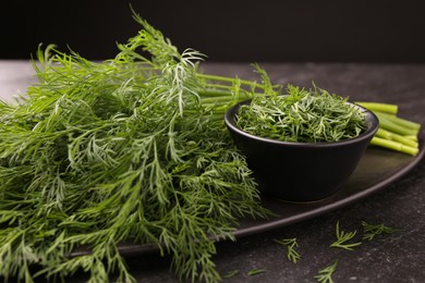 Photo of Sprigs of fresh dill and bowl with cut one on dark textured table, closeup