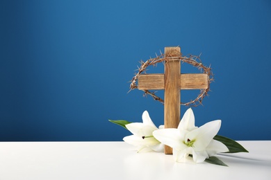 Photo of Wooden cross, crown of thorns and blossom lilies on table against color background, space for text