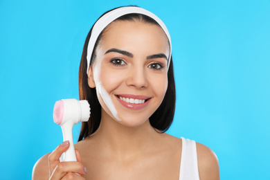 Photo of Young woman using facial cleansing brush on light blue background. Washing accessory
