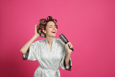 Photo of Beautiful young woman in silk bathrobe with hair curlers singing into hairbrush on pink background. Space for text