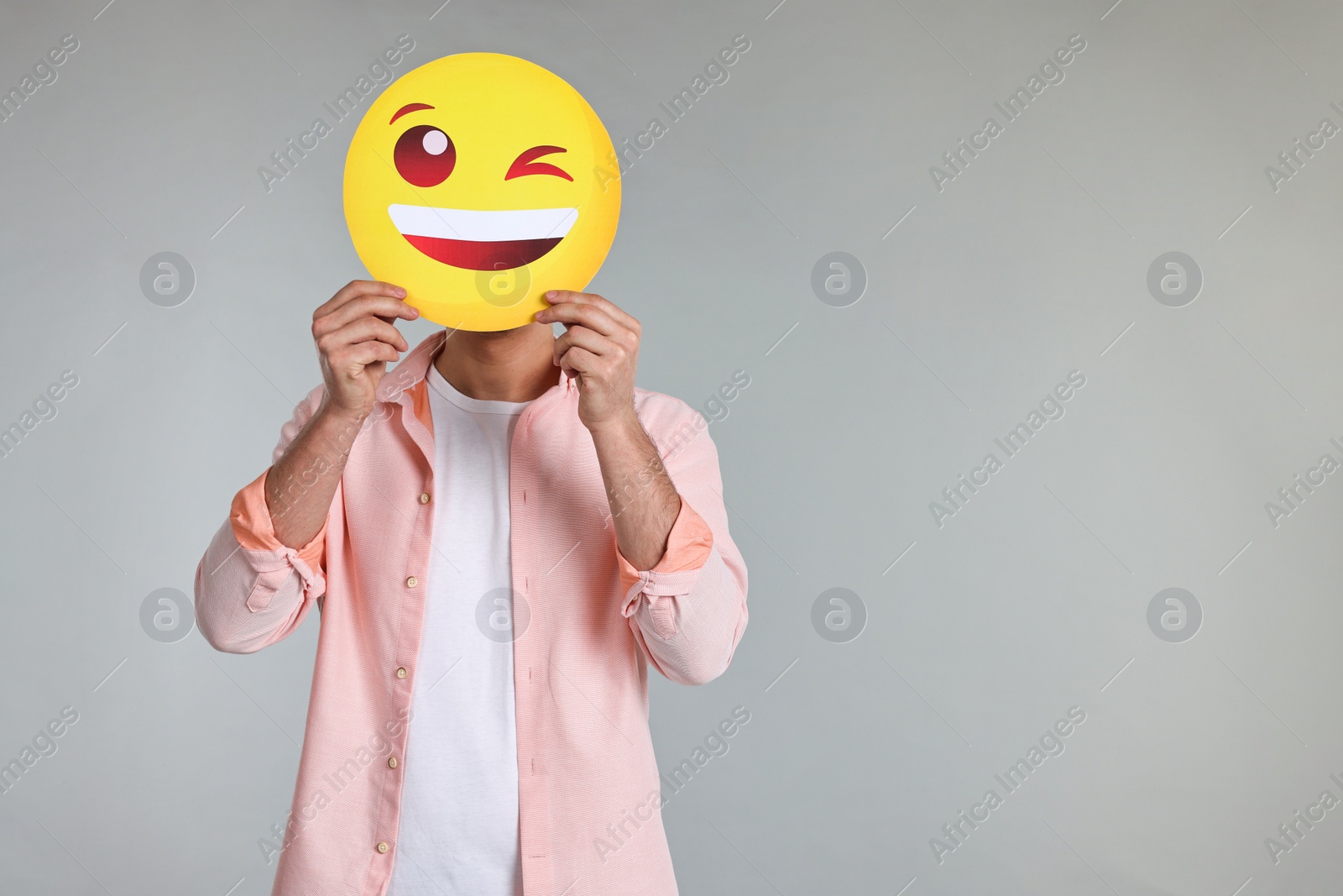 Photo of Man covering face with happy winking emoticon on grey background. Space for text