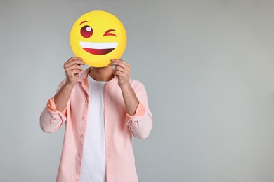 Photo of Man covering face with happy winking emoticon on grey background. Space for text