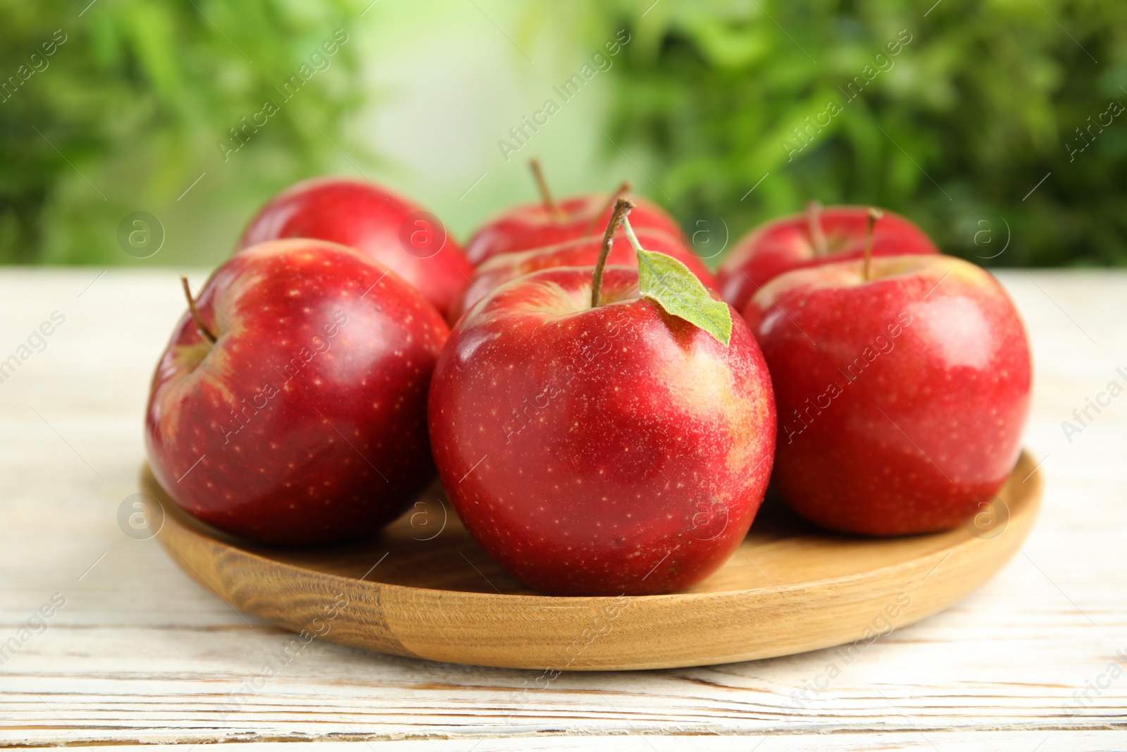 Photo of Wooden plate with ripe juicy red apples on white table against blurred background