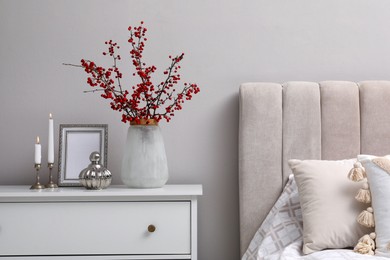 Photo of Hawthorn branches with red berries, candles and frame on chest of drawers in bedroom