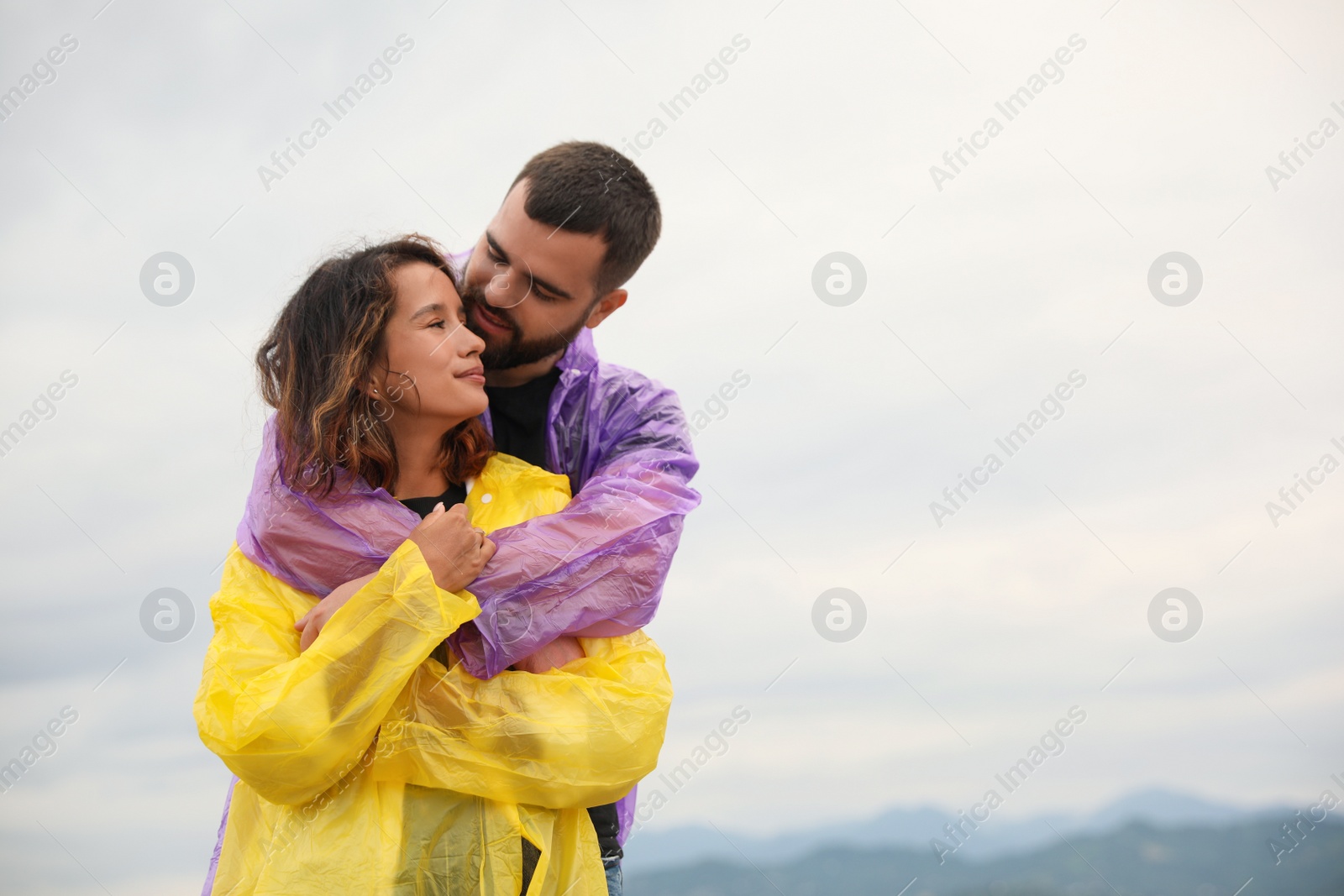 Photo of Young couple in raincoats enjoying time together under rain on beach, space for text