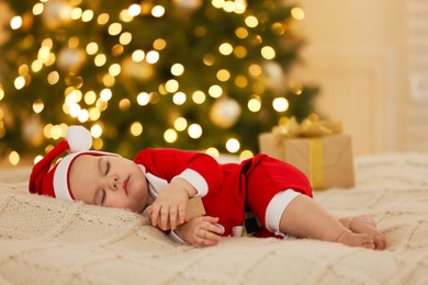 Cute baby in Christmas costume sleeping on knitted blanket against blurred festive lights. Winter holiday