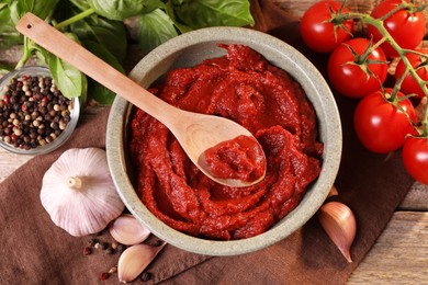 Photo of Bowl of tasty tomato paste with spoon and ingredients on wooden table, flat lay