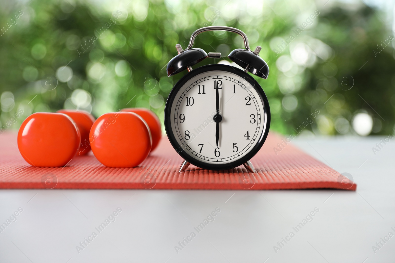 Photo of Alarm clock, dumbbells and yoga mat on grey table against blurred background. Morning exercise