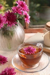 Photo of Beautiful chrysanthemum flowers and cup of tea on beige textured table near window. Autumn still life