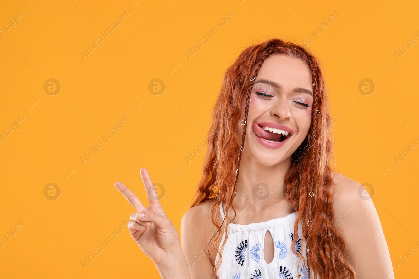 Photo of Beautiful young hippie woman showing her tongue and V-sign on orange background, space for text