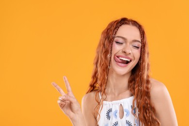 Beautiful young hippie woman showing her tongue and V-sign on orange background, space for text