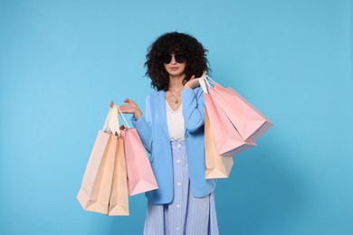 Happy young woman with shopping bags and stylish sunglasses on light blue background