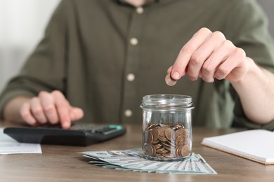 Photo of Financial savings. Man putting coin into glass jar while using calculator at wooden table, closeup