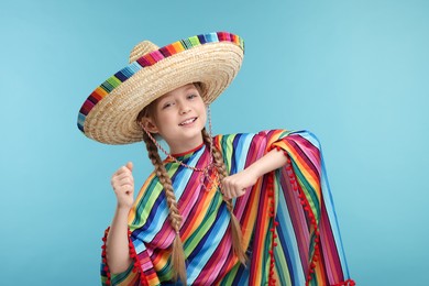 Cute girl in Mexican sombrero hat and poncho dancing on light blue background