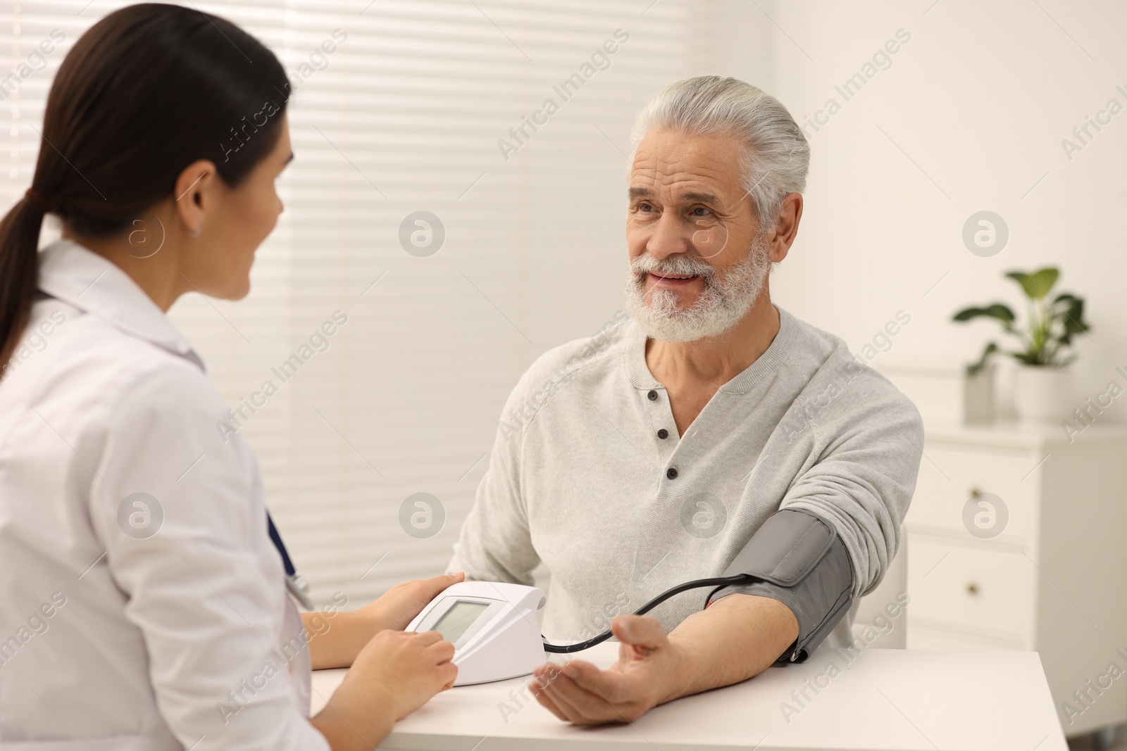 Photo of Nurse measuring elderly patient's blood pressure at white table in hospital