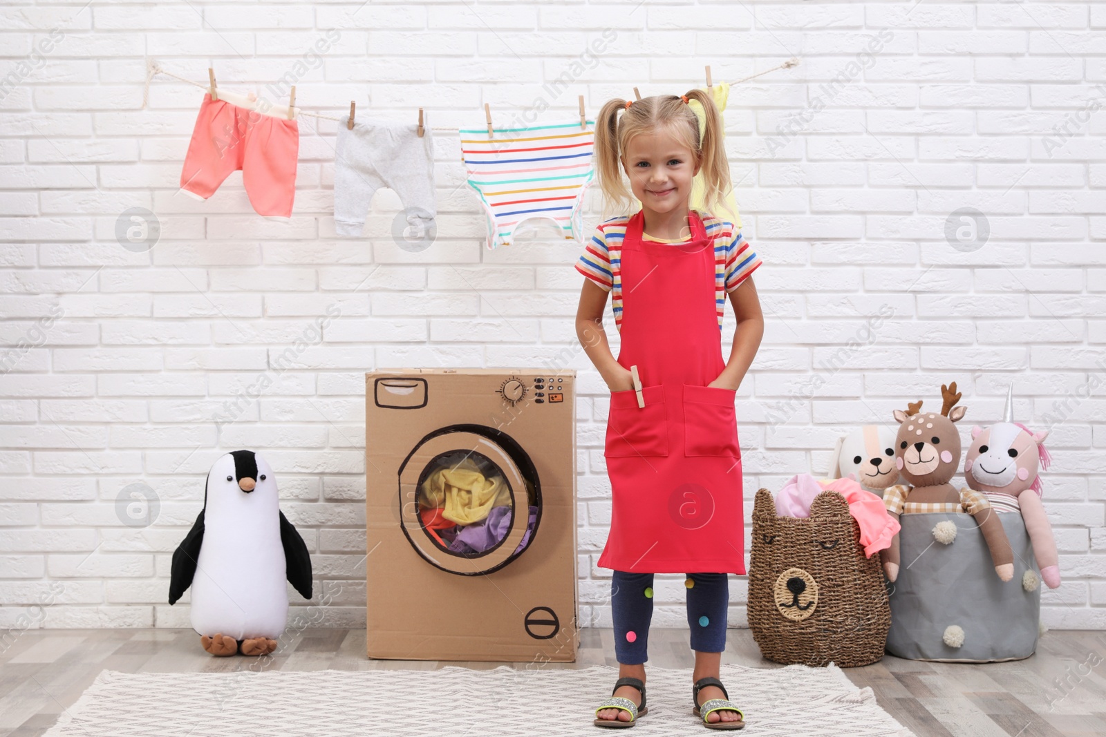 Photo of Little girl playing with toy cardboard washing machine indoors